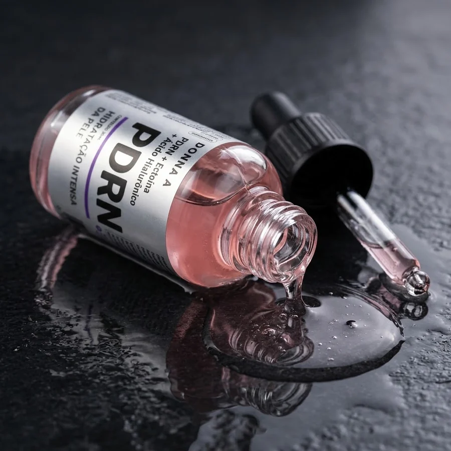 Frasco do sérum PDRN deitado, com o líquido rosa escorrendo e formando uma poça
